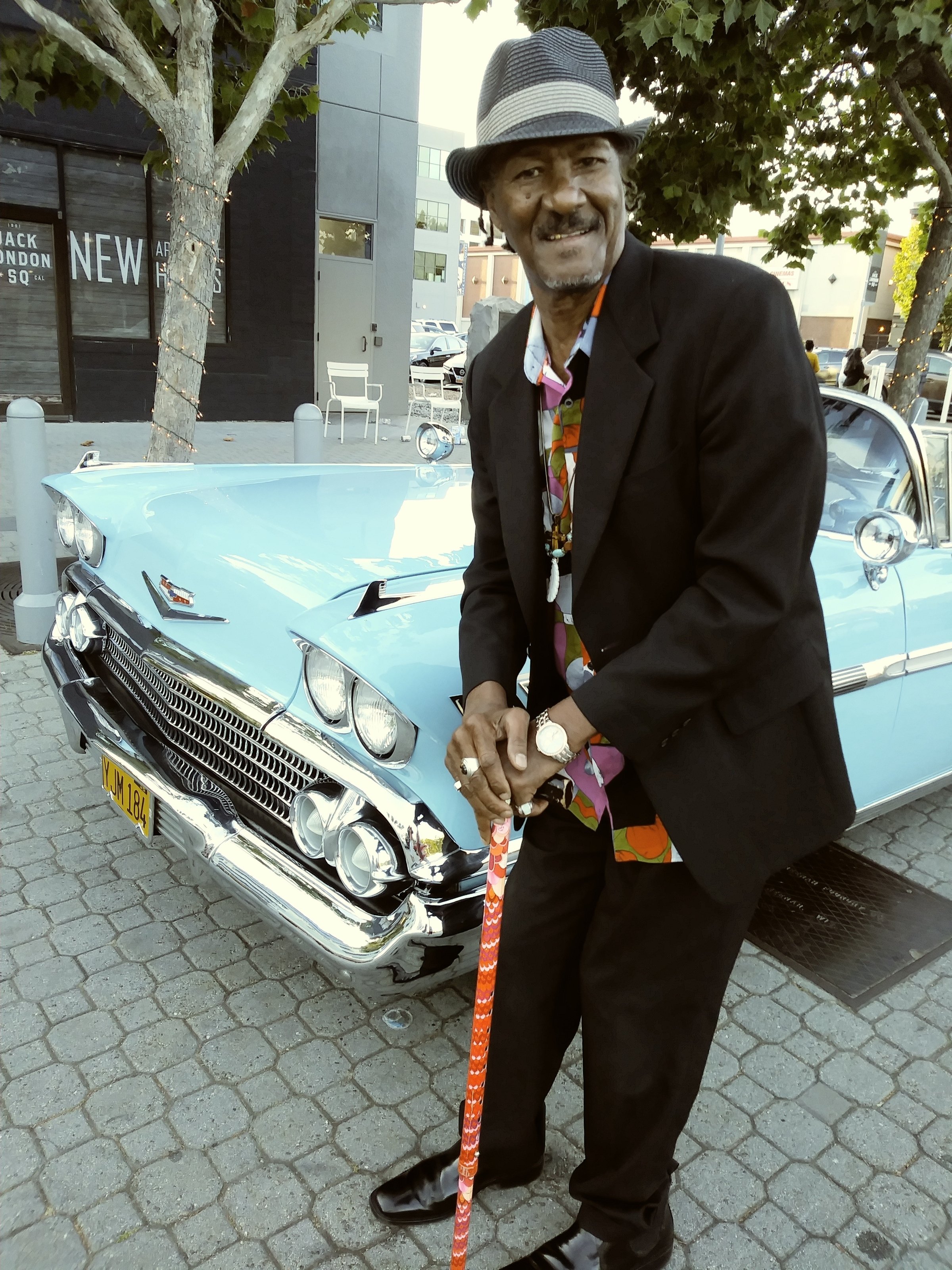 Fillmore Slim in a dark suit and hat, leaning on a cane beside a light blue vintage Chevrolet